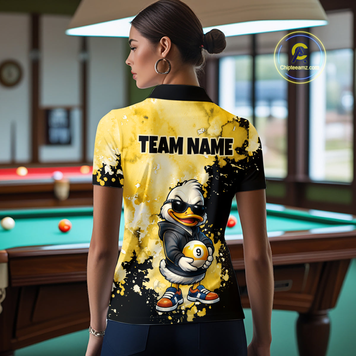 A woman stands in a pool room wearing a short-sleeve collared polo shirt from the back. The shirt is bright yellow and black with a cartoon duck mascot graphic holding a billiard ball and the text 'TEAM NAME' across the upper back.