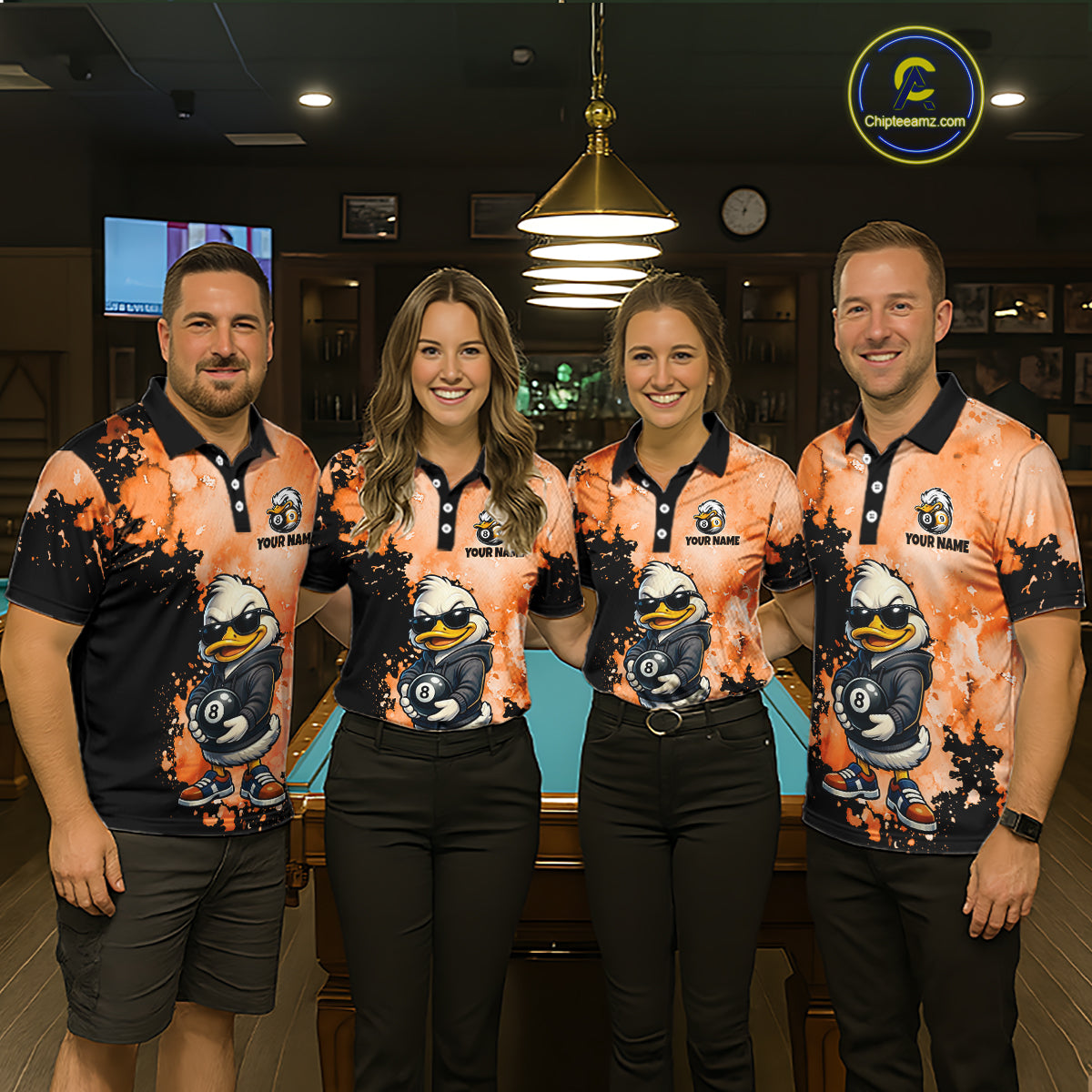 Four people standing together in a dimly lit indoor venue, wearing matching orange and black polo shirts with a cartoon duck graphic and the text 'Troy Jackson'. A pool table is visible behind them