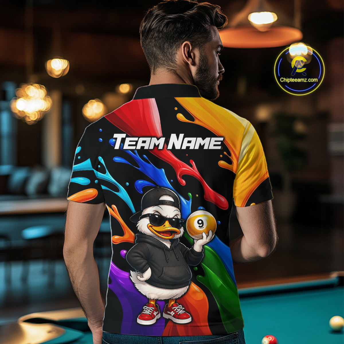 A man viewed from behind wearing a short-sleeve collared shirt with a colorful rainbow paint-splatter design, a cartoon duck character holding a 9-ball, and the text 'TEAM NAME' on the back. The background shows a blurred pool hall with a green pool table.
