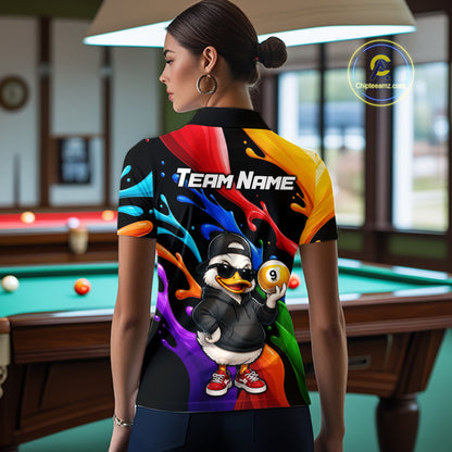 A woman stands in a billiards room wearing a custom short-sleeve polo shirt from the back. The shirt features a colorful paint splatter background with a cartoon duck wearing a black cap and sunglasses, holding a 9-ball, and the text 'TEAM NAME' across the upper back.