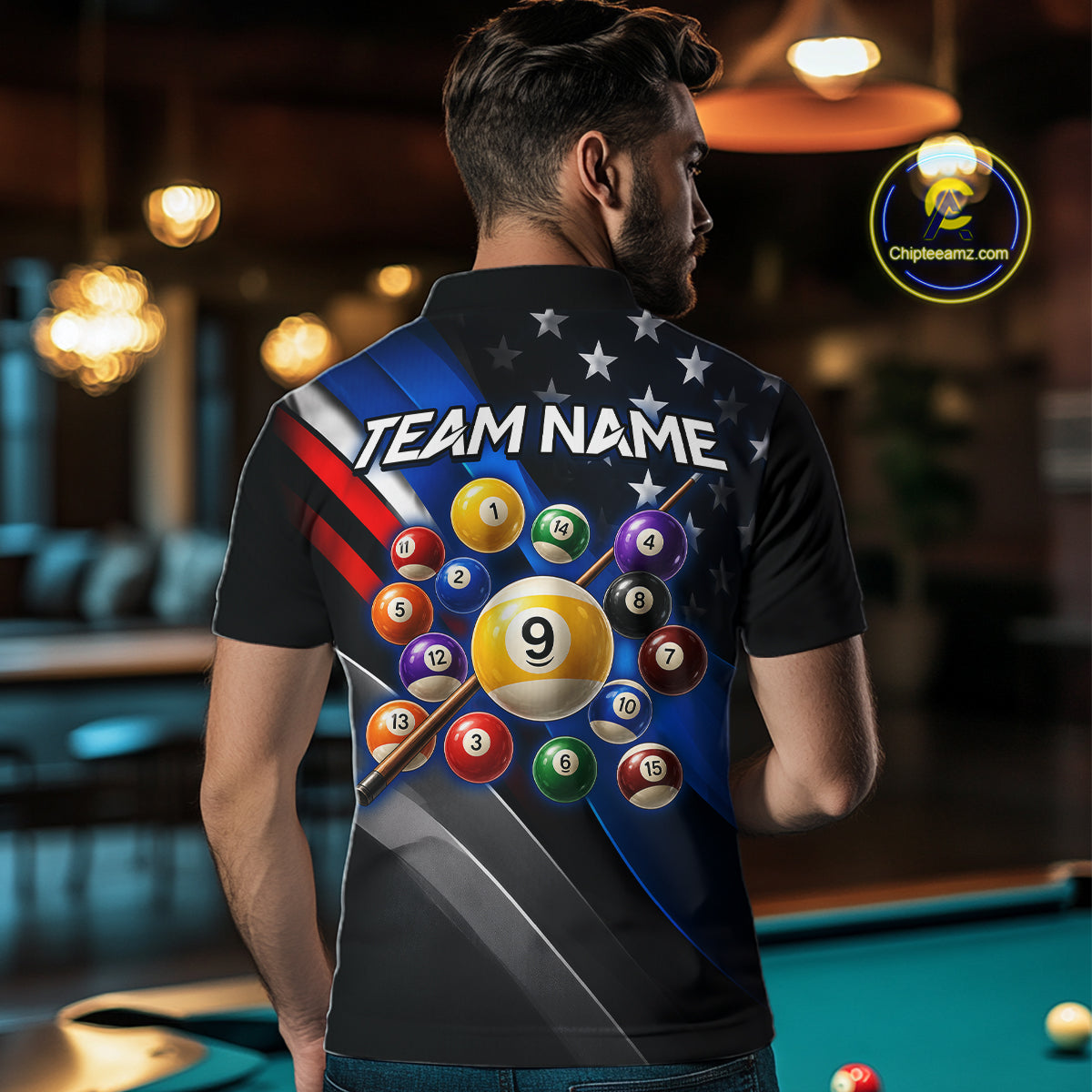 A man viewed from behind wearing a custom black short-sleeve polo shirt with a large back graphic featuring billiards balls, stars, stripes, and the text 'TEAM NAME'. The background shows a blurred pool hall environment with a green pool table and warm hanging lights.