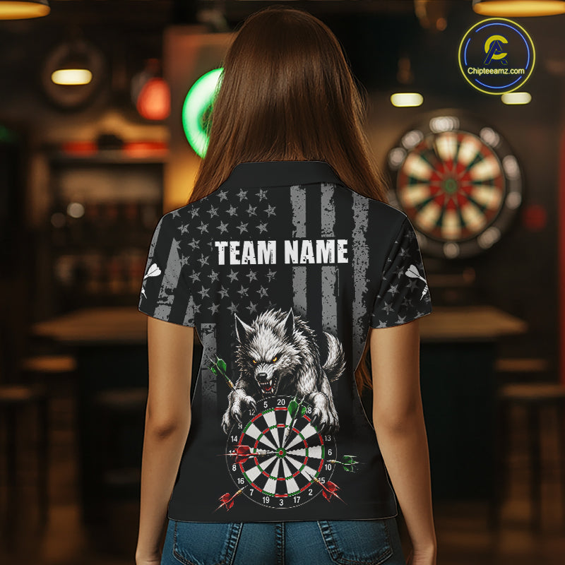 A woman viewed from behind wearing a black short-sleeve polo shirt with a large back graphic featuring a white wolf, a dartboard, and distressed American flag elements. The shirt has placeholder text reading 'TEAM NAME' and is shown in a dimly lit bar environment with a dartboard visible in the background.