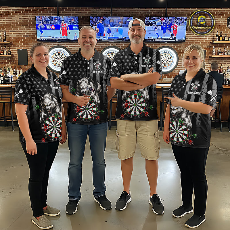 Four people standing together in a bar wearing matching black short-sleeve polo shirts with a white star pattern, a large dartboard graphic on the front. The background shows a brick wall, bar shelves, and mounted sports screens.