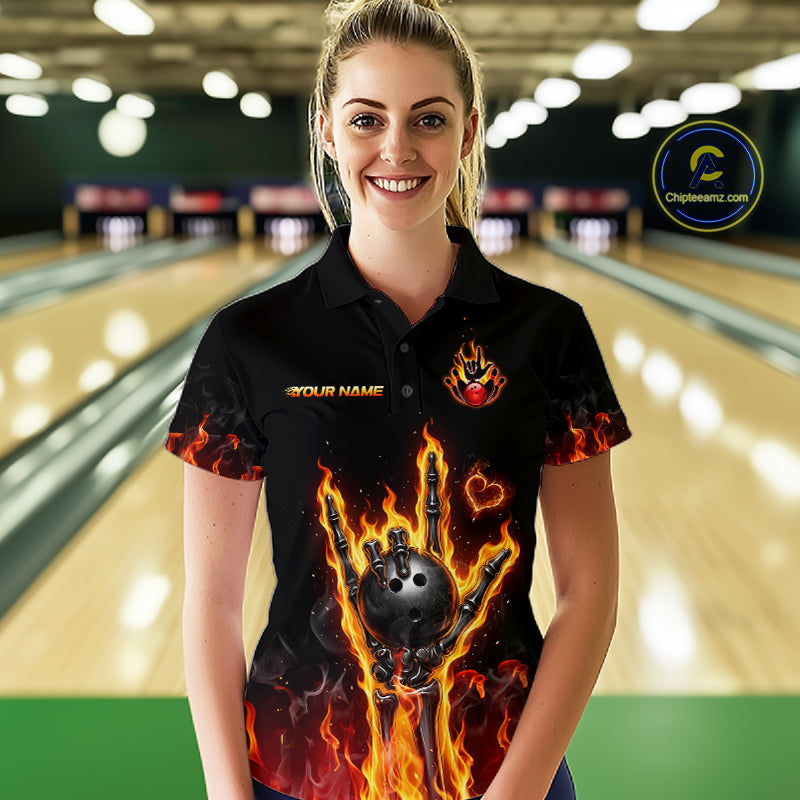A woman wearing a black short-sleeve bowling-themed polo shirt with a flaming bowling ball graphic and flame details, standing in a bowling alley. The shirt features a customizable name print on the left chest and a flame logo on the right chest.