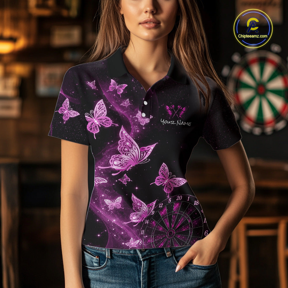 A woman wearing a black short-sleeve polo shirt with a purple and pink butterfly print, custom text on the chest, and a dartboard graphic near the hem, set against a blurred indoor background with a dartboard.