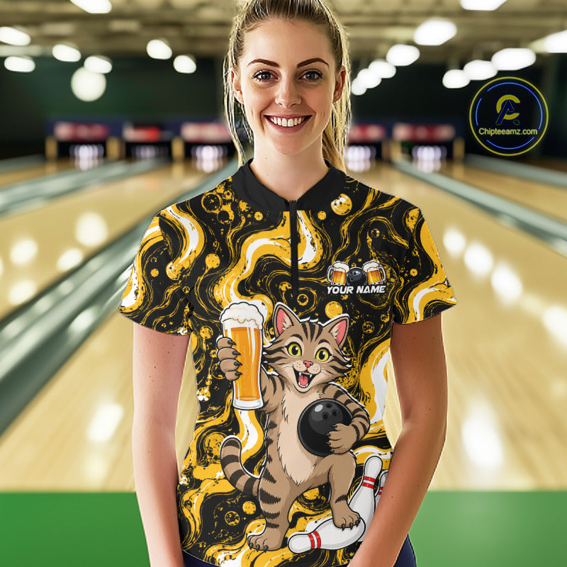 A woman standing in a bowling alley wearing a short-sleeve bowling shirt with a black and yellow abstract swirl pattern and a cartoon cat holding a bowling ball and beer glass. The shirt has a black collar and a partial placket, with 'YOUR NAME' printed on the chest.