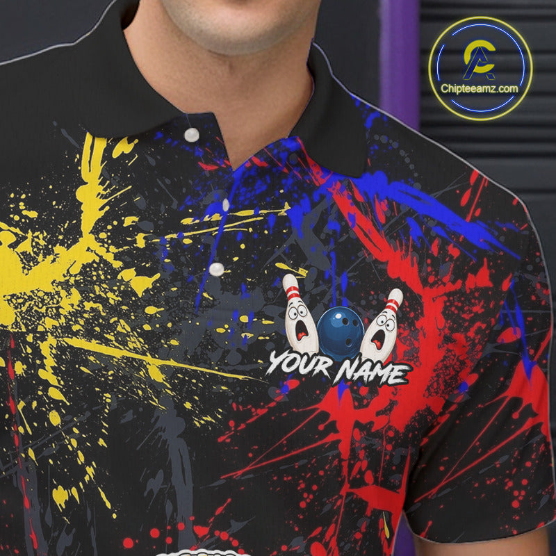 A close-up of a black short-sleeve polo shirt with a colorful bowling-themed graphic print. The design features red, blue, and yellow paint splatters on a black background, with two cartoon bowling pins and a blue bowling ball near the center, and the text 'YOUR NAME' below.