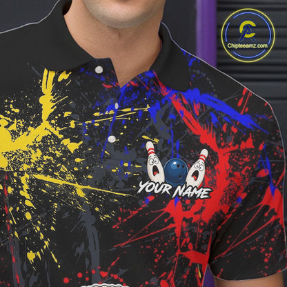 A close-up of a black short-sleeve polo shirt with a colorful bowling-themed graphic print. The design features red, blue, and yellow paint splatters on a black background, with two cartoon bowling pins and a blue bowling ball near the center, and the text 'YOUR NAME' below.