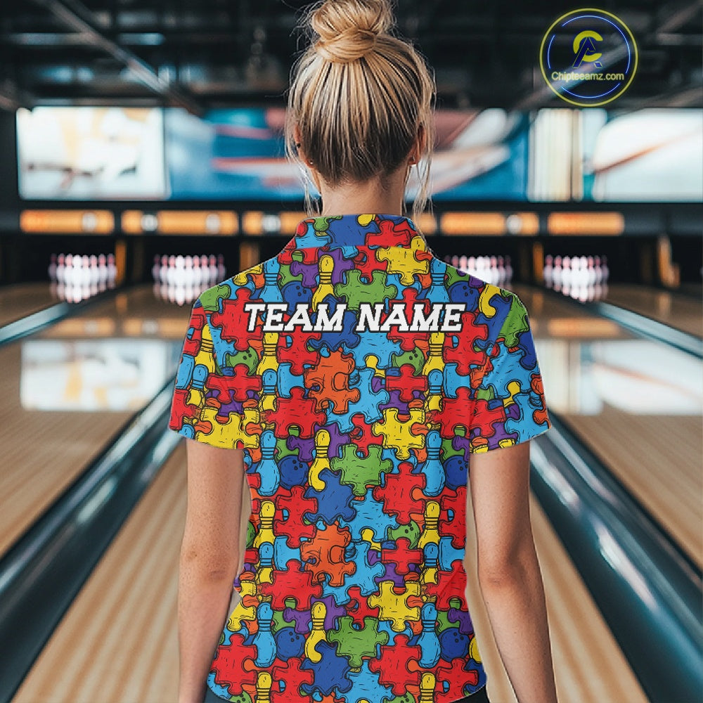 A woman viewed from behind wearing a short-sleeve shirt covered in a colorful puzzle-piece pattern, standing in a bowling alley. The shirt has the words 'TEAM NAME' printed across the back.