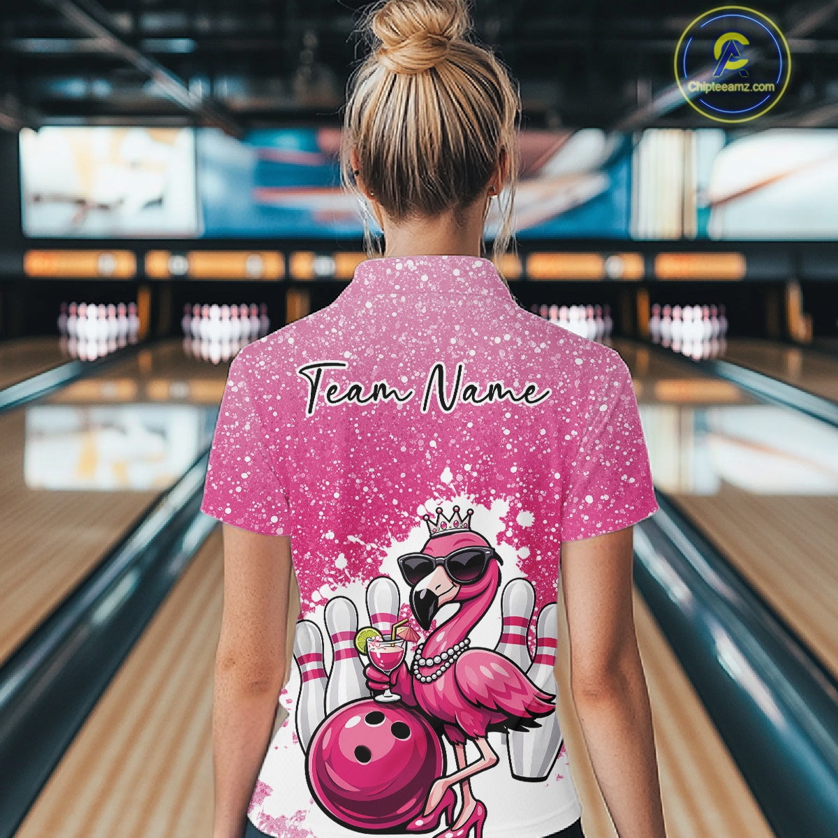 Back view of a female model in a bowling center wearing a pink quarter-zip shirt featuring a crowned flamingo holding a cocktail beside a bowling ball.