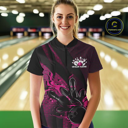 Female model at a bowling lane wearing a black and pink quarter zip shirt, front view showcasing flames bowling ball knocking down pins with bold geometric styling