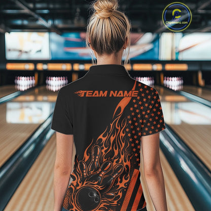 Back view of a female model in a bowling center wearing a black polo shirt featuring an orange flaming bowling ball and patriotic pin artwork.