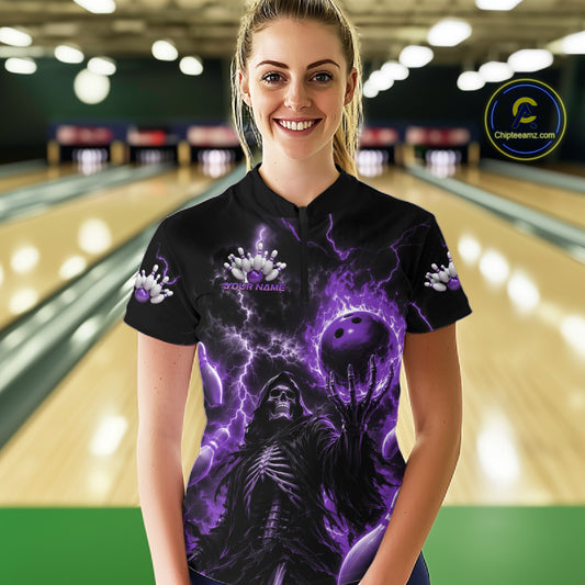 A woman stands in a bowling alley wearing a black short-sleeve quarter-zip shirt with a dramatic purple and black graphic print featuring a hooded figure, lightning, and a bowling ball. 