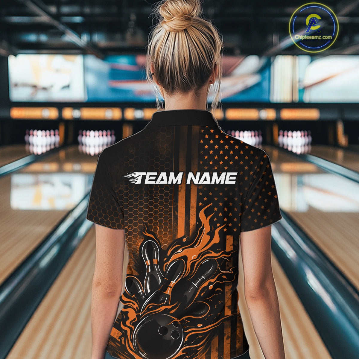 Back view of a female model in a bowling center wearing a black and orange quarter-zip shirt featuring a bowling strike scene with flame effects.